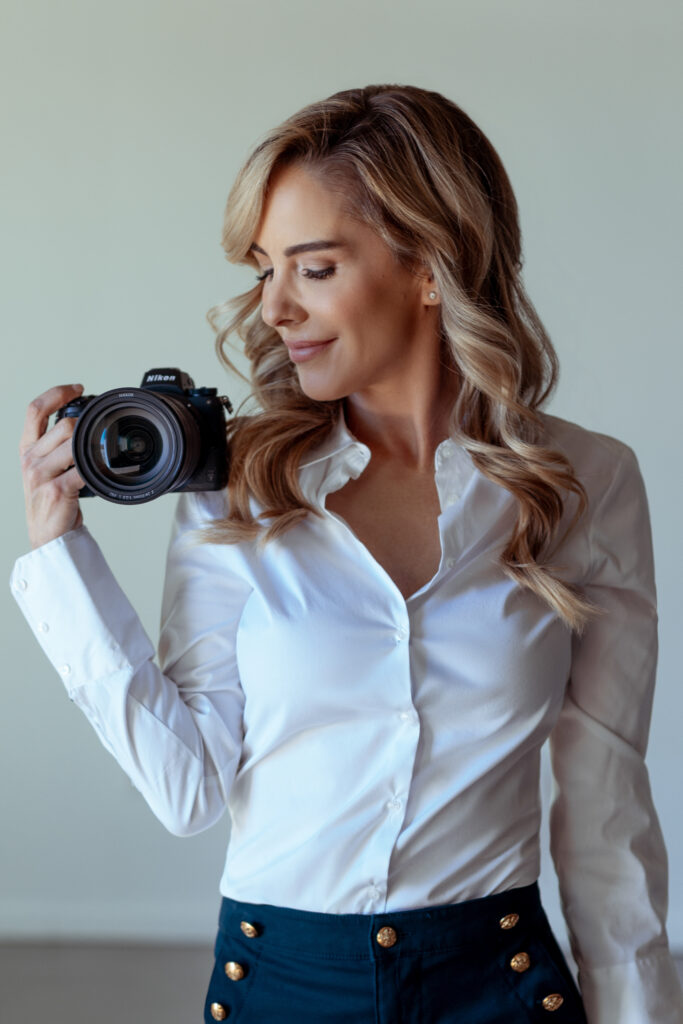 Woman in white blouse holding a Nikon camera, looking down with wavy blonde hair